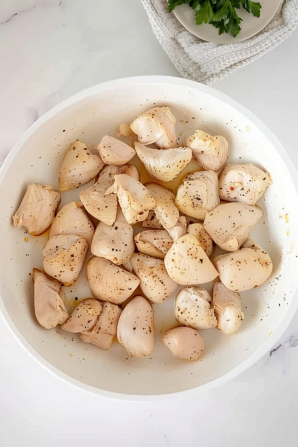 A white bowl of chicken chunks on a white marble countertop.