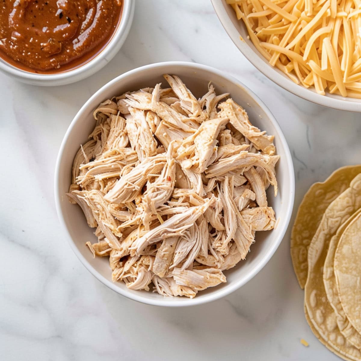 Corn tortillas with bowls of shredded chicken, enchilada sauce and shredded cheese.