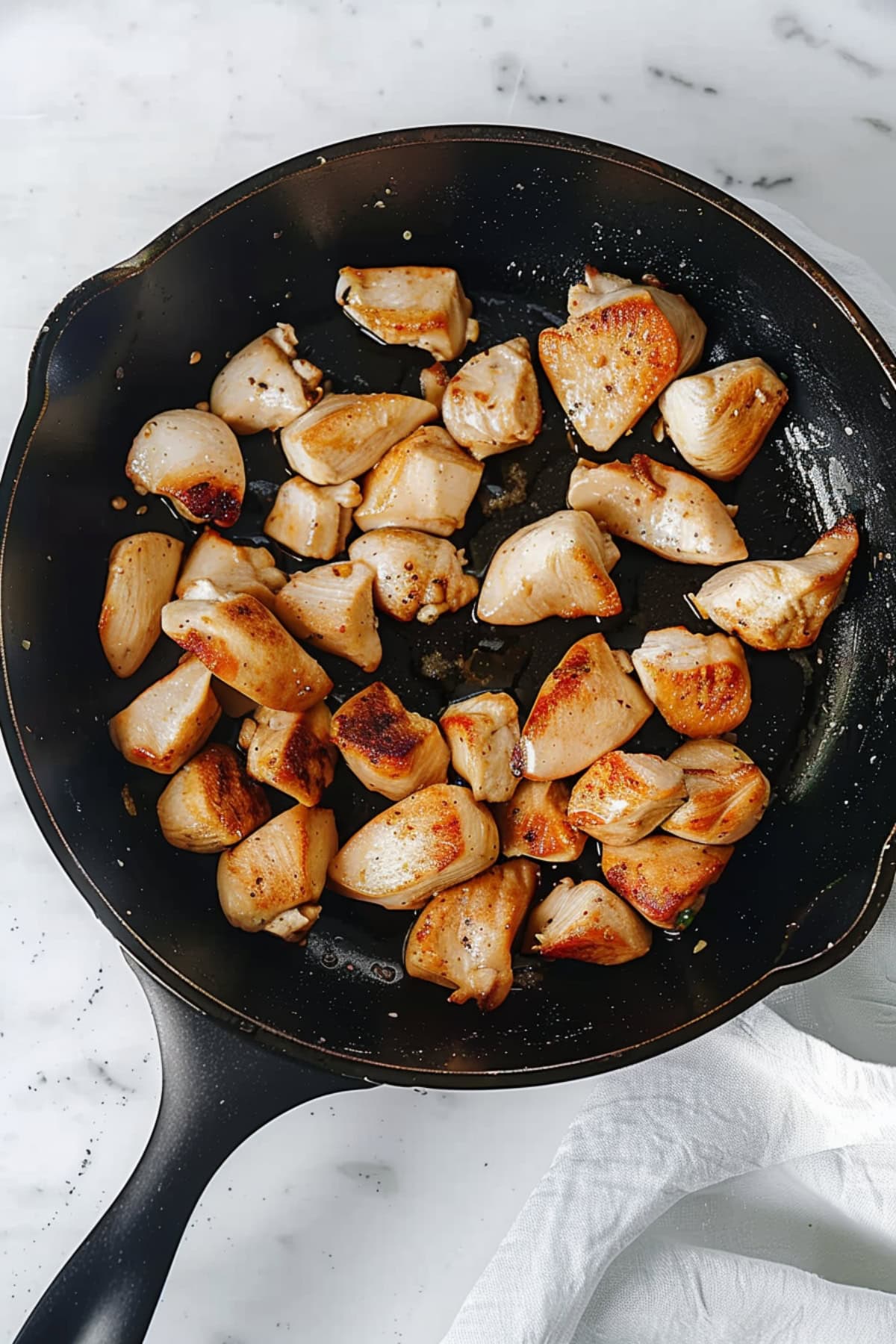 A black skillet of cooked chicken pieces on a white marble table.