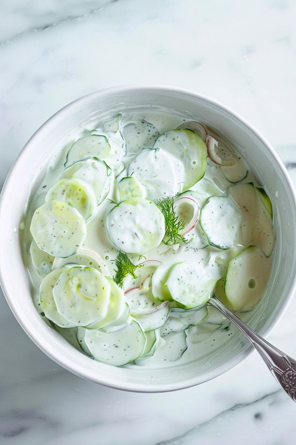 Bowl of creamy cucumber salad in a white bowl on a white marble table.