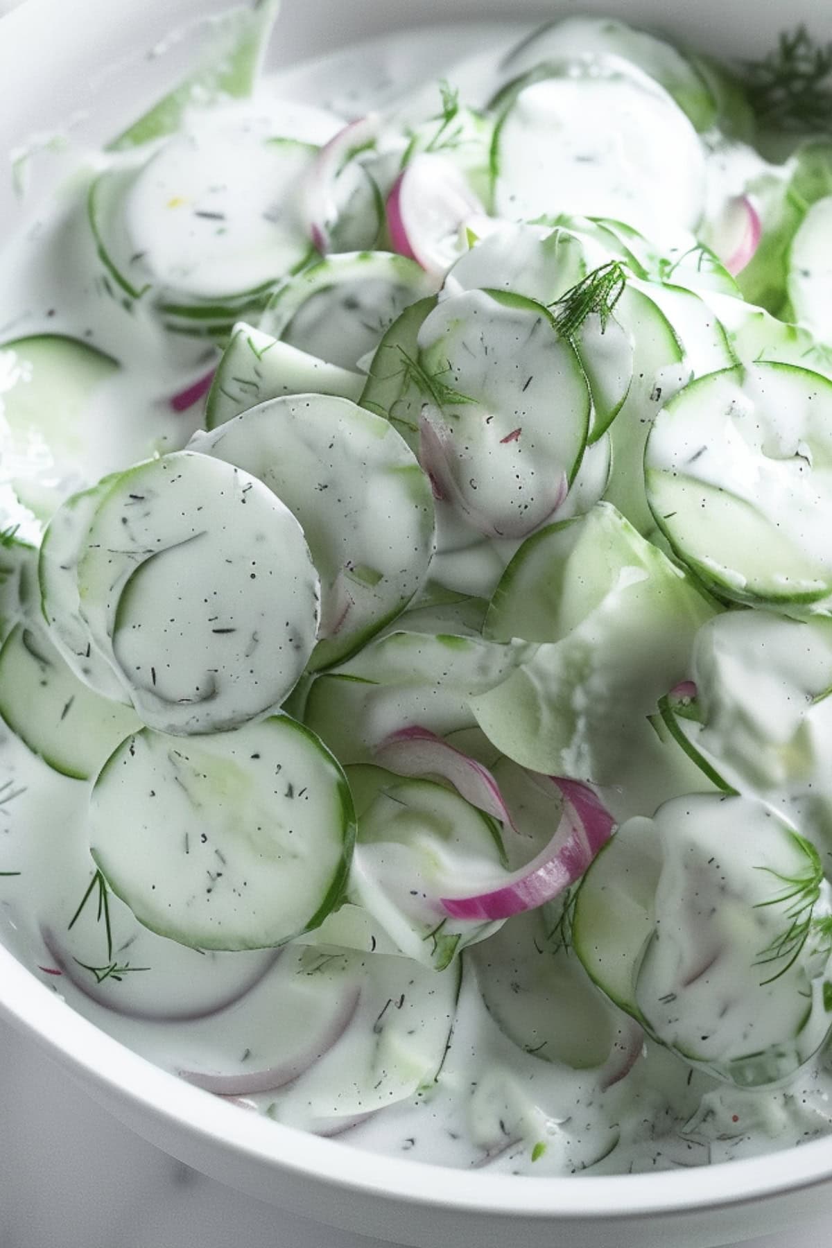 Thinly sliced cucumber, onions and chopped dills with creamy dressing in a white bowl.