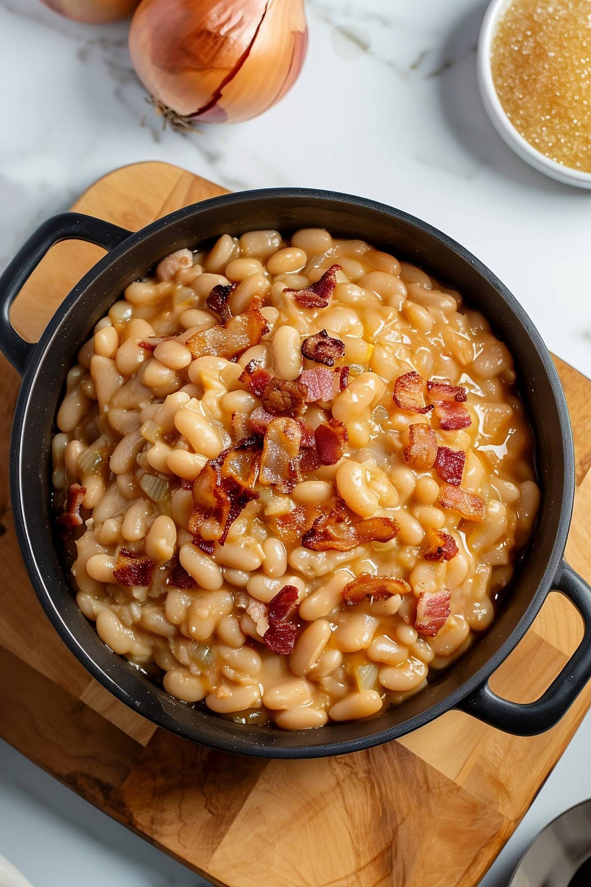 Top View of Grandma Brown's Baked Beans with Bacon in a Cast Iron Pan on a Wooden Cutting Board with Onions and Sugar Around the Pan