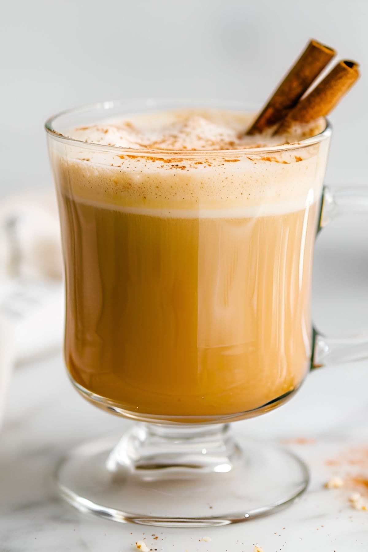 Close Up of Hot Buttered Rum in a Glass with a Foamy Top, Cinnamon Powder, and Two Cinnamon Sticks for Garnish