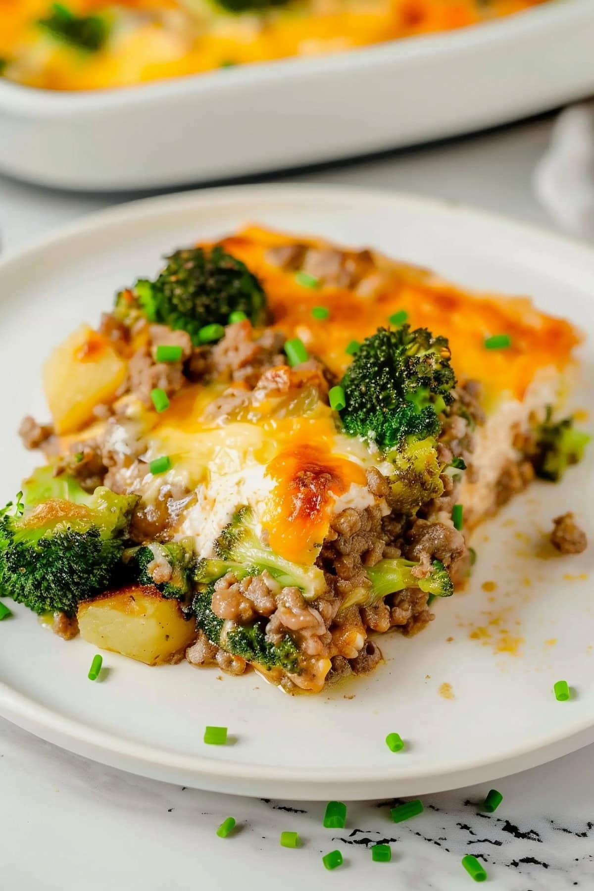 Close up of a Slice of Italian Sausage Breakfast Casserole with Ground Italian Sausage, Broccoli, Potatoes, and Lots of Cheese on a Plate with the Remaining Casserole in the Dish in the Background