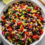 Close Up Top View of Mexican Bean Salad with Black Beans, White Beans, Peppers, Tomatoes, Corn, Red Onion, and Cilantro in a Bowl on a White Marble Table with Wooden Spoons and Limes