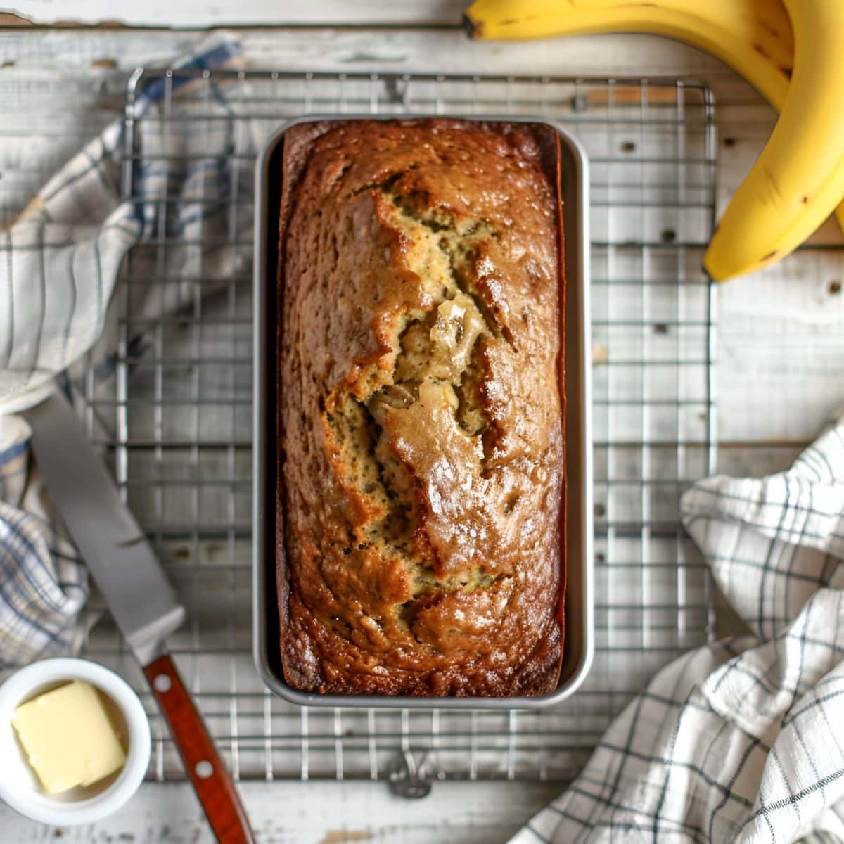 Top View of a Loaf of Freshly Baked Paula Deen Banana Bread in a Loaf Tin, Cooling on a Wire Rack with Fresh Bananas, Ramekin of Butter, a Knife, and a Kitchen Towel to the Side