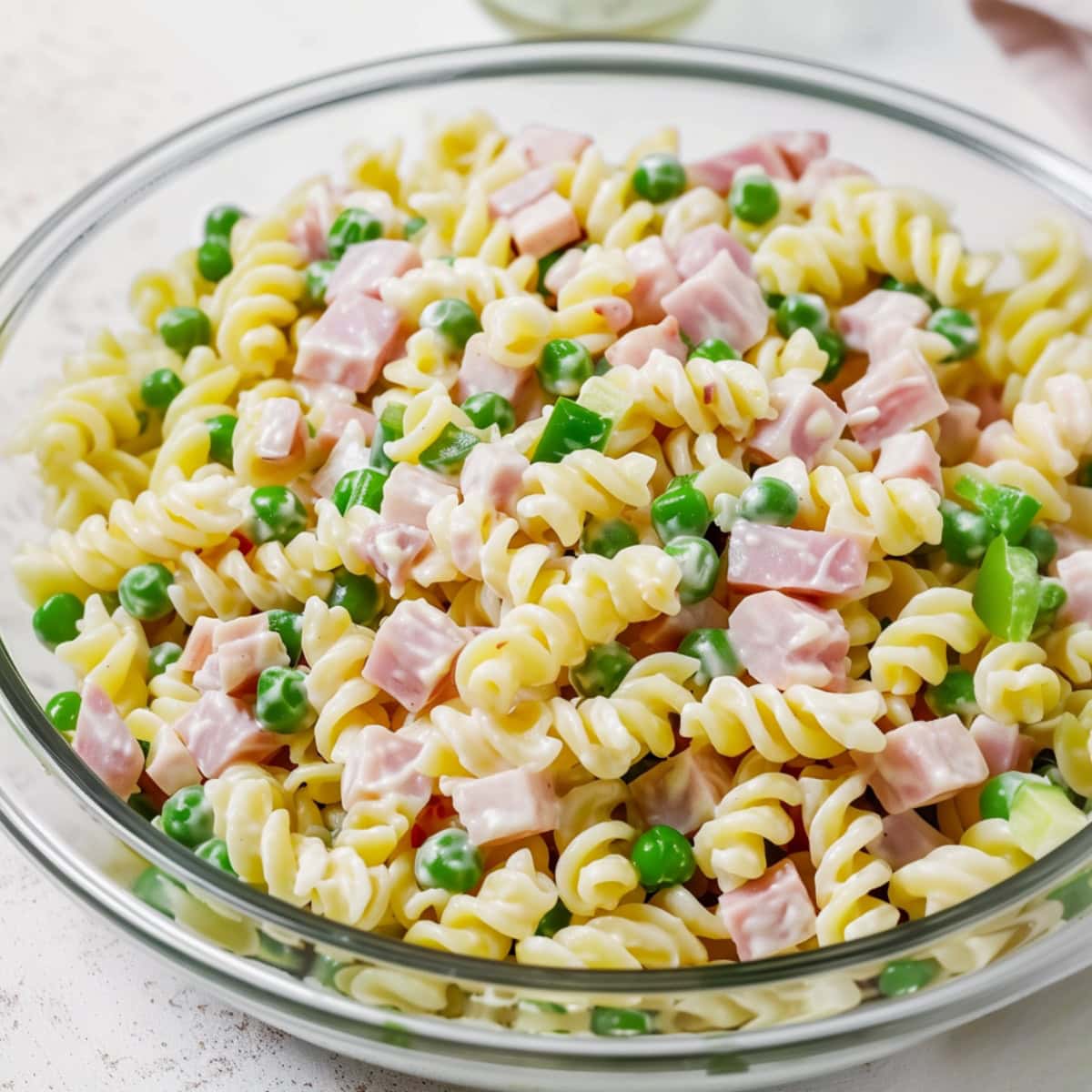 Bowl of rotini pasta, diced ham, frozen peas, and diced green pepper covered in dressing.