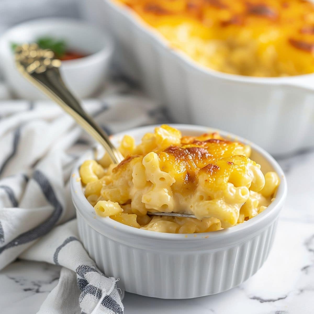 Gooey, Cheesy Sweetie Pie's Mac and Cheese with a Browned Cheese Topping and a Spoon in a White Bowl on a White Marble Table and a Full Casserole Dish in the Background
