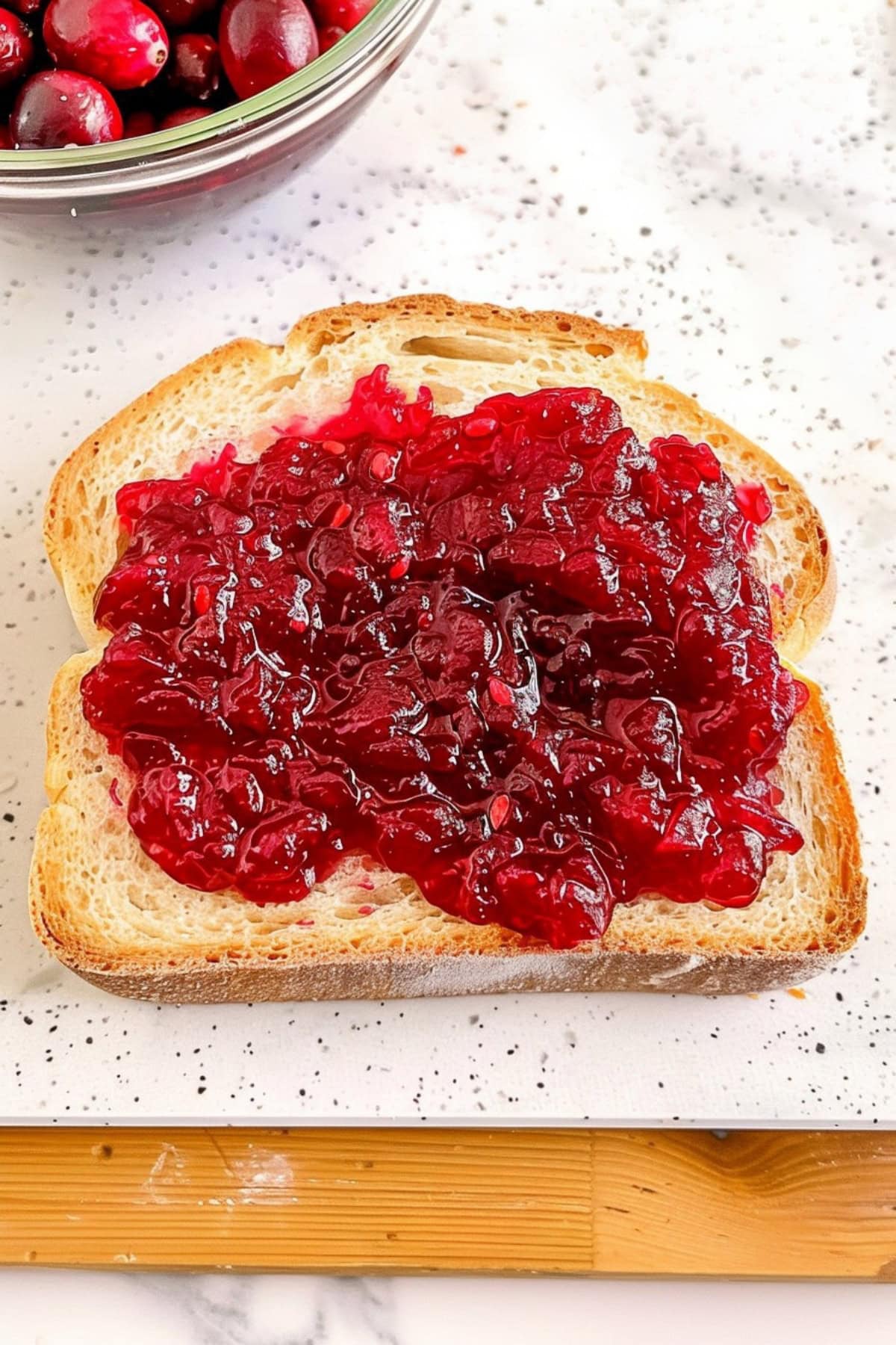 A slice of white bread with cranberry sauce spread over the entire surface. The light gray background features an empty wooden board underneath. A bowl filled with cranberries can be seen in one corner. 