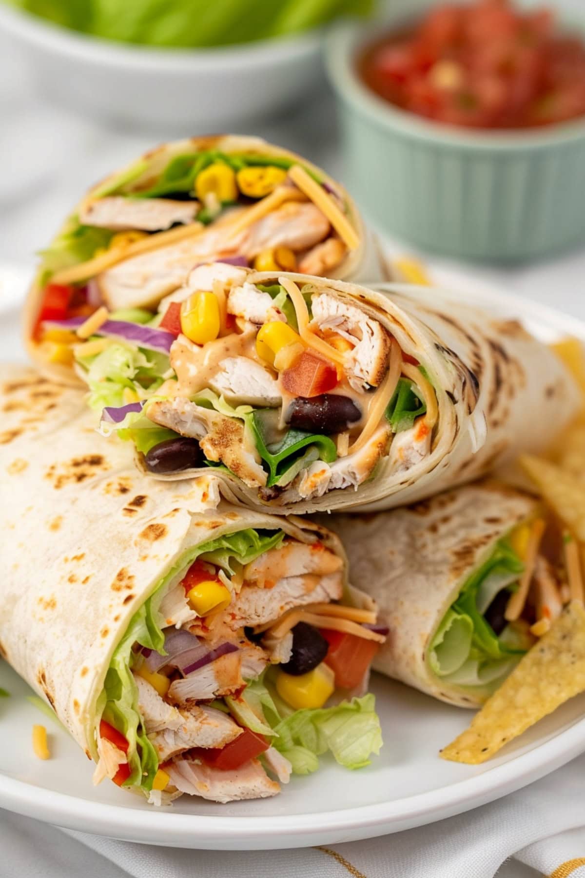 Chicken wrap with chicken, lettuce, black beans, corn, red bell pepper, onion, cilantro, tortilla strips and cheese with ranch sauce wrapped in flour tortilla arranged in a white plate. 