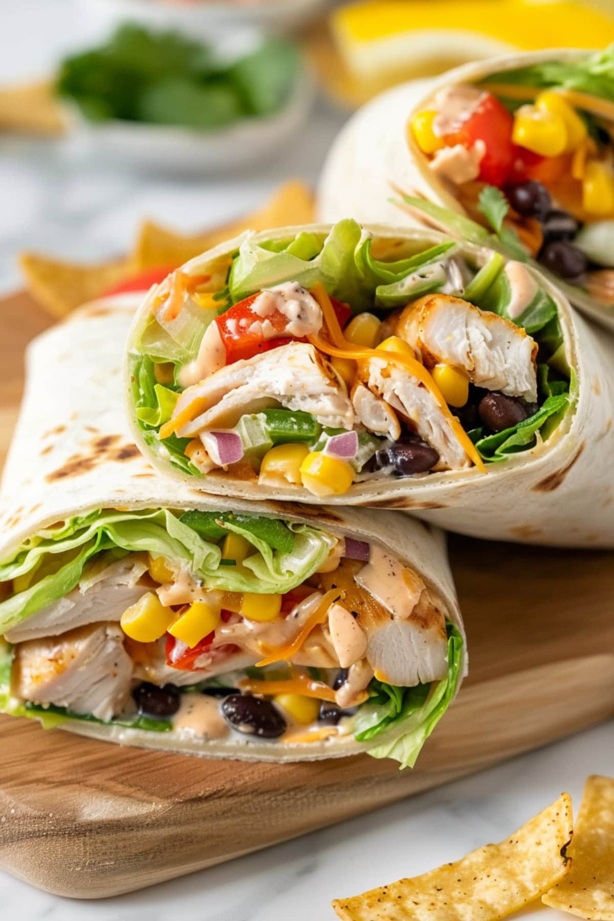 Sliced in half chicken wrap with sliced chicken, lettuce, black beans, corn, red bell pepper, onion, cilantro, tortilla strips and cheese with ranch sauce wrapped in flour tortilla arranged in a wooden board.
