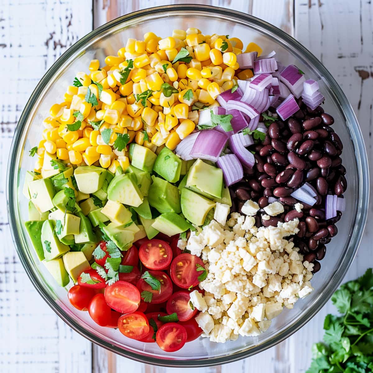 Black beans, corn kernels, red bell pepper diced, diced red onion, cherry tomatoes, jalapeno pepper minced, fresh cilantro chopped, avocado diced, crumbled feta cheese in a glass bowl on a white wooden table.