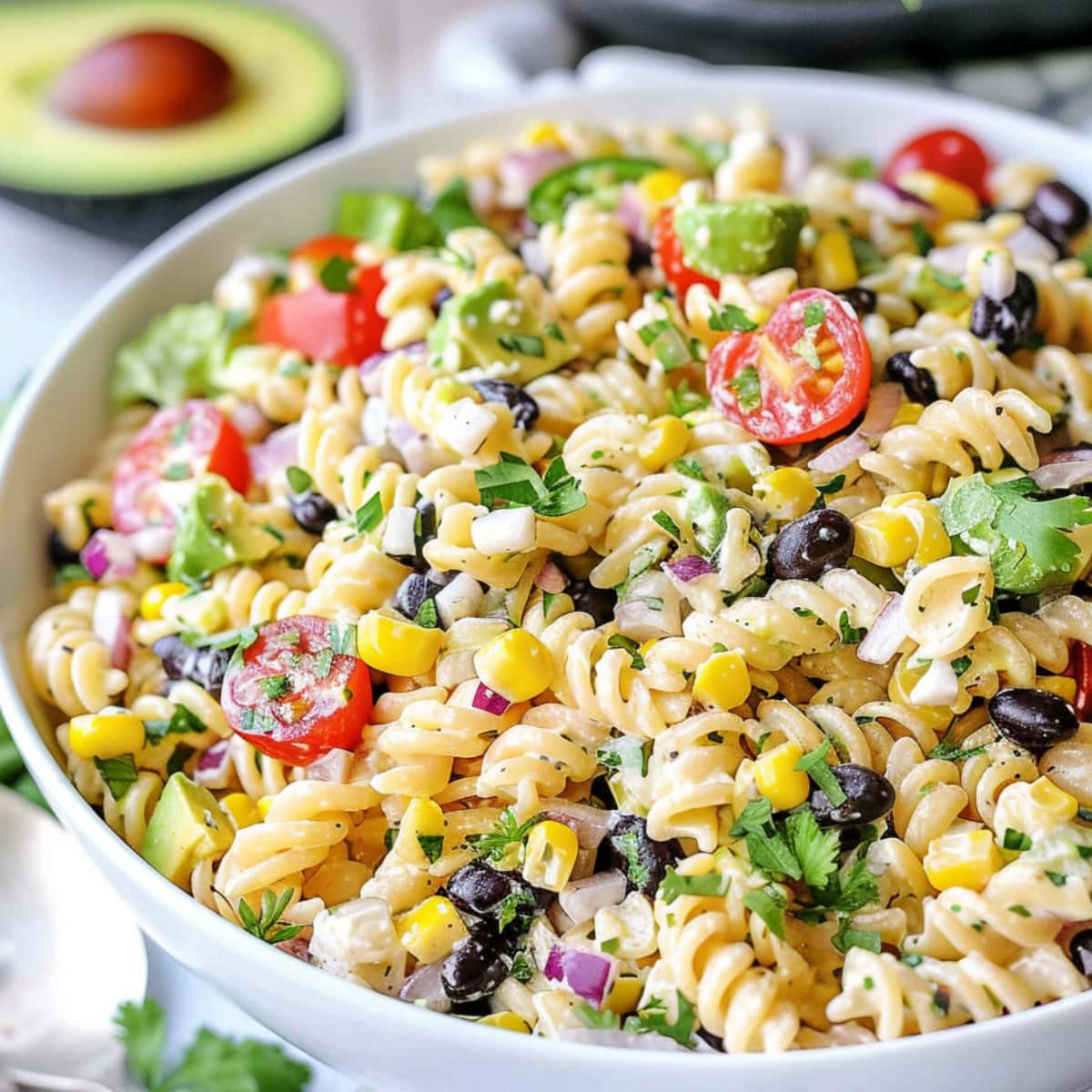 Rotini pasta with black beans, corn kernels, red bell pepper diced, diced red onion, cherry tomatoes, jalapeno pepper minced, fresh cilantro chopped, avocado diced, crumbled feta cheese coated in a creamy white dressing served on a white bowl on a white wooden table.
