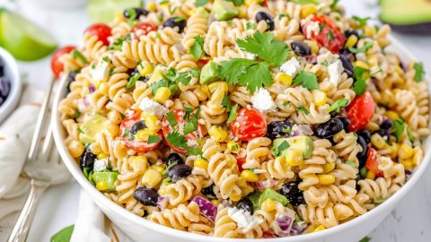 Southwest pasta salad made with black beans, corn kernels, red bell pepper diced, diced red onion, cherry tomatoes, jalapeno pepper minced, fresh cilantro chopped, avocado diced, crumbled feta cheese coated in a creamy white dressing served on a white bowl on a white wooden table.