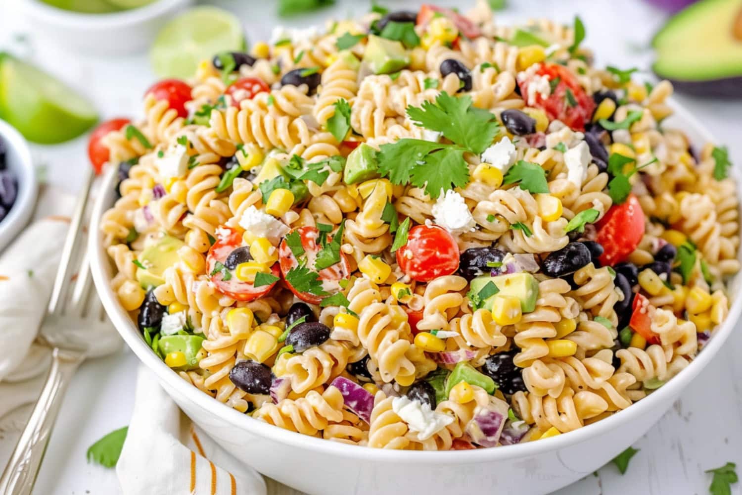 Southwest pasta salad made with black beans, corn kernels, red bell pepper diced, diced red onion, cherry tomatoes, jalapeno pepper minced, fresh cilantro chopped, avocado diced, crumbled feta cheese coated in a creamy white dressing served on a white bowl on a white wooden table.