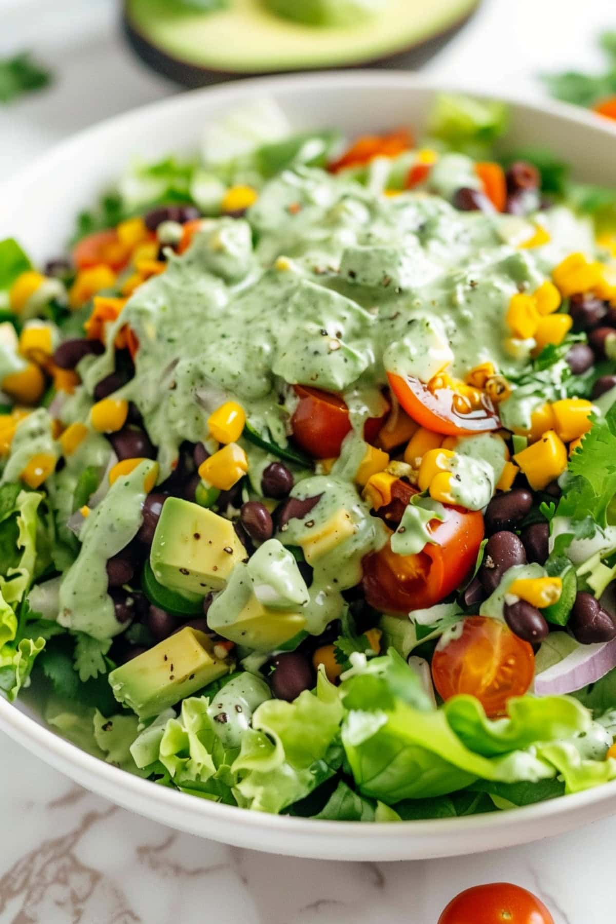 Romaine, hearty black beans, sweet corn, crunchy veggies, salty cotija, and creamy avocado with avocado dressing served on a white plate.