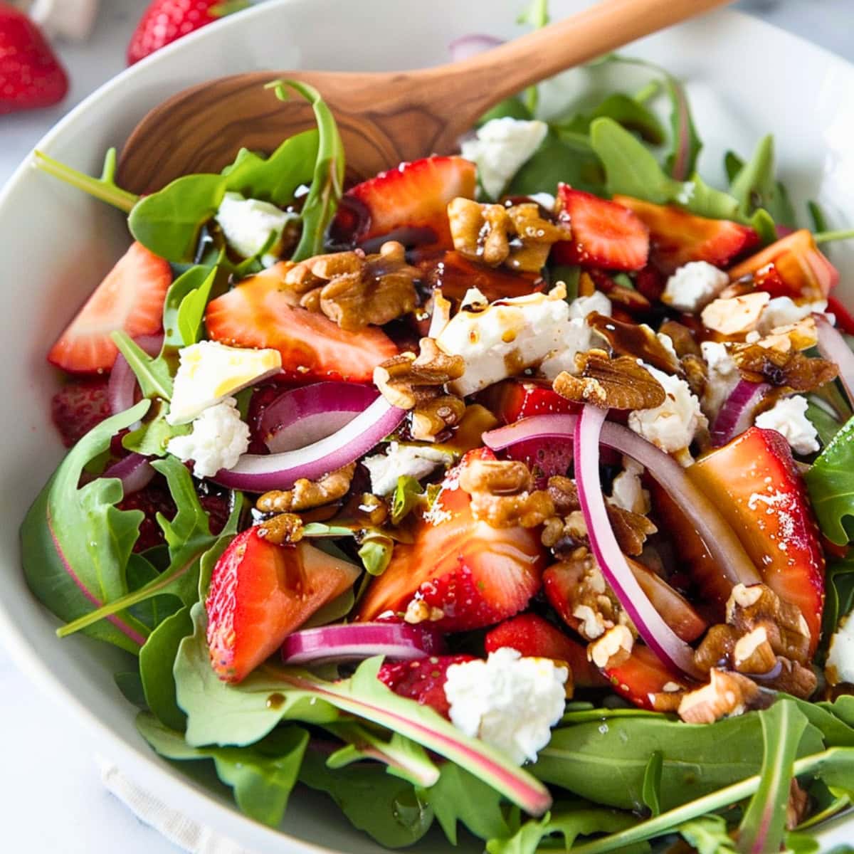 Mixed greens, sliced strawberries, red onion, and toasted walnuts with goat cheese in a white bowl.