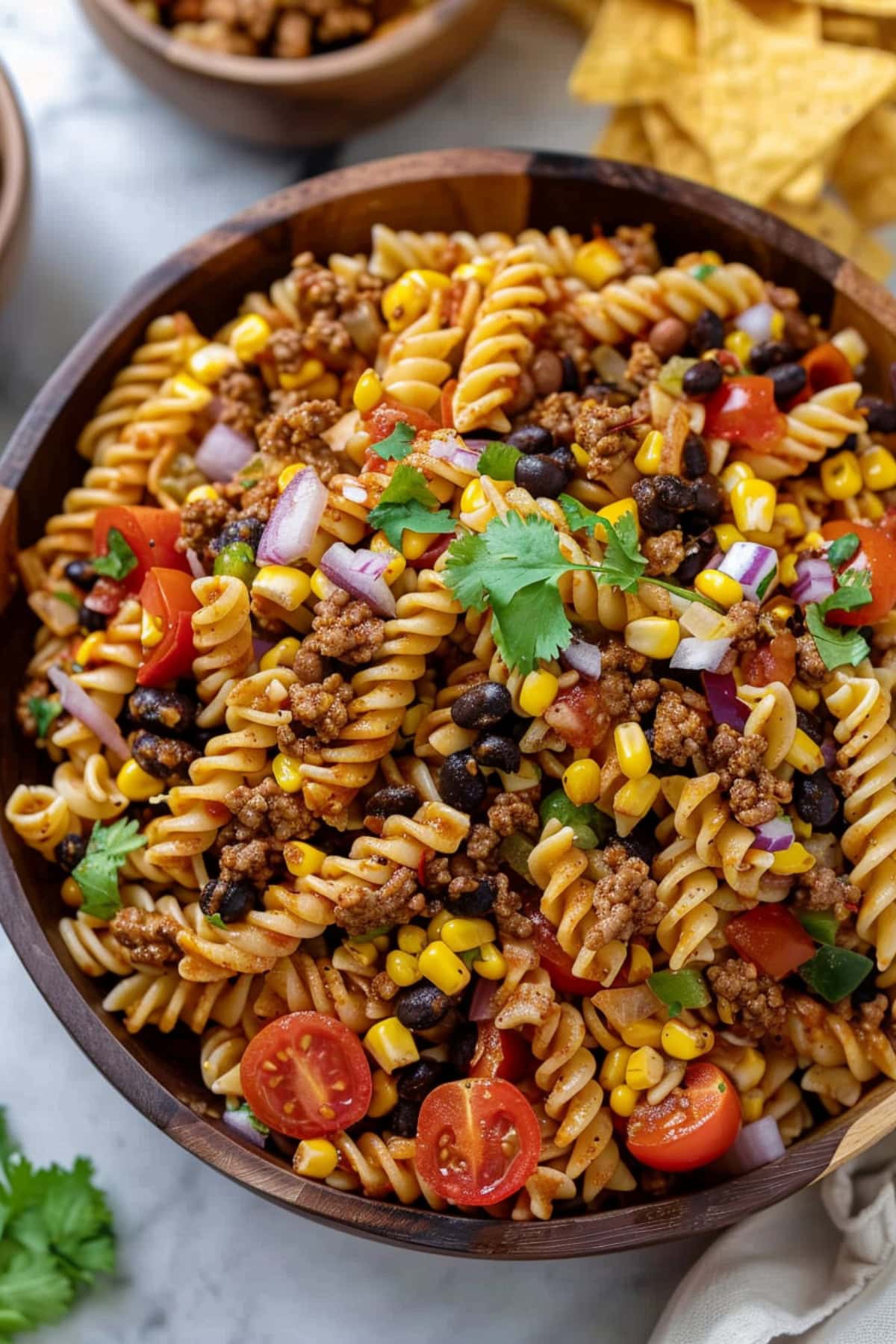 Taco pasta salad with tomatoes, red onions, black beans, corn kernels and cilantro on a wooden bowl with tortilla cchips on the side.