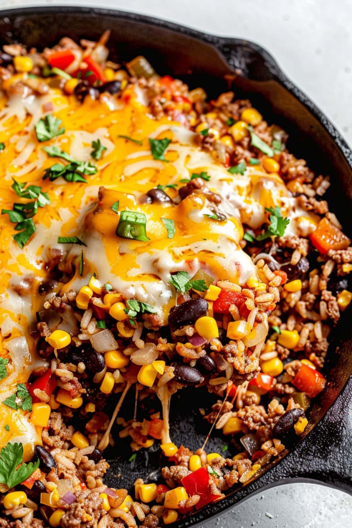 Ground beef with diced red bell pepper, diced red onion, corn kernels, cooked rice, black beans, salsa topped with melted cheese on a cast iron skillet pan on a white marble table.