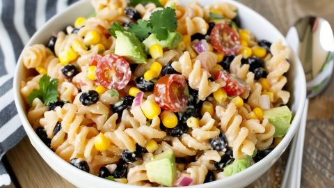 A festive presentation of Tex-Mex pasta salad in a large serving bowl, featuring black beans, corn, avocado and cherry tomatoes.