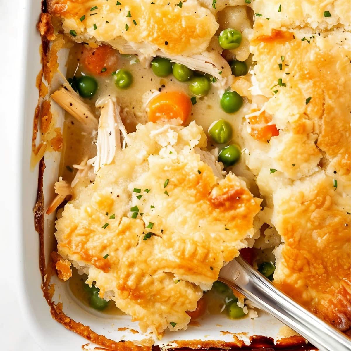 Close Top View of TikTok Chicken Cobbler- Shredded Chicken, Peas, Carrots, Sauce, and Flaky Biscuit Topping- in a Casserole Dish with a Serving Utensil