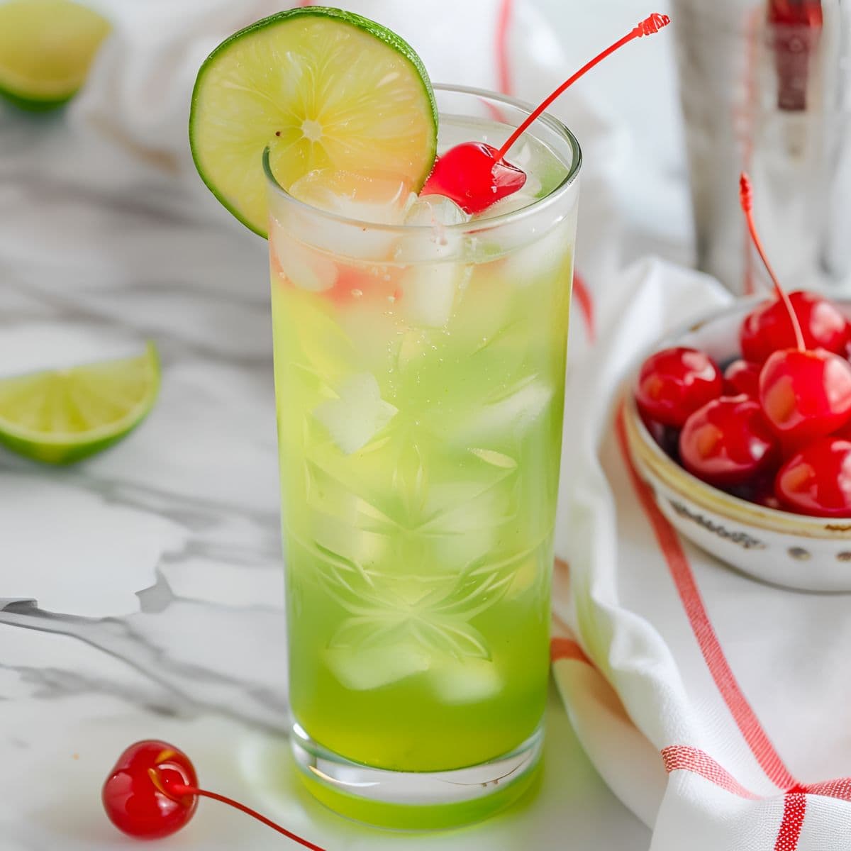 Close Up of Tokyo Tea in a Tall Glass with Ice, a Maraschino Cherry Garnish, and a Lime Wheel with Cherries and Limes in the Background