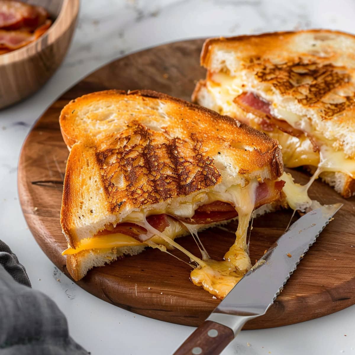 Grilled cheese sandwich cut in half featuring crispy bacon and melting cheese on a chopping board with a serrated knife