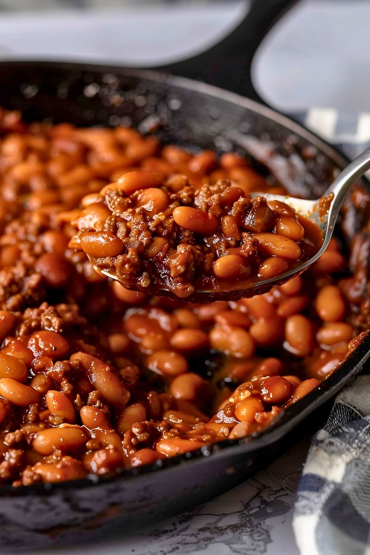Close Up of a Spoonful of BBQ Baked Beans- with Ground Beef, Beans, and Sauce- Held Over a Cast Iron Skillet with More BBQ Baked Beans