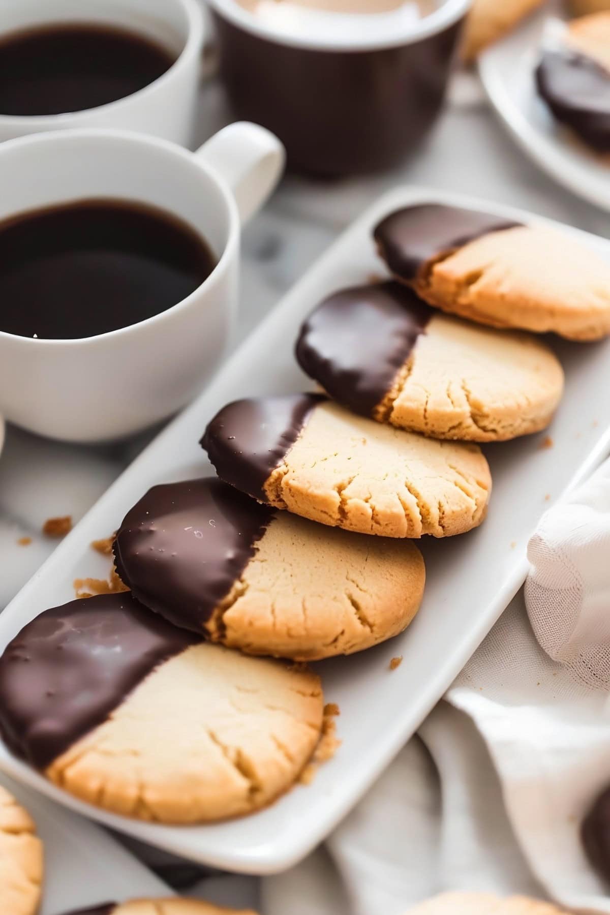 Chocolate-Dipped Shortbread Cookies Lined Up on a Thin Serving Plate with Cups of Coffee in the Background