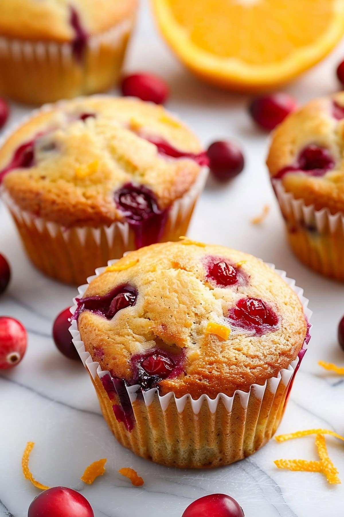 Close Up of Four Cranberry Orange Muffins on a White Marble Table with Fresh Cranberries and Oranges Scattered Around