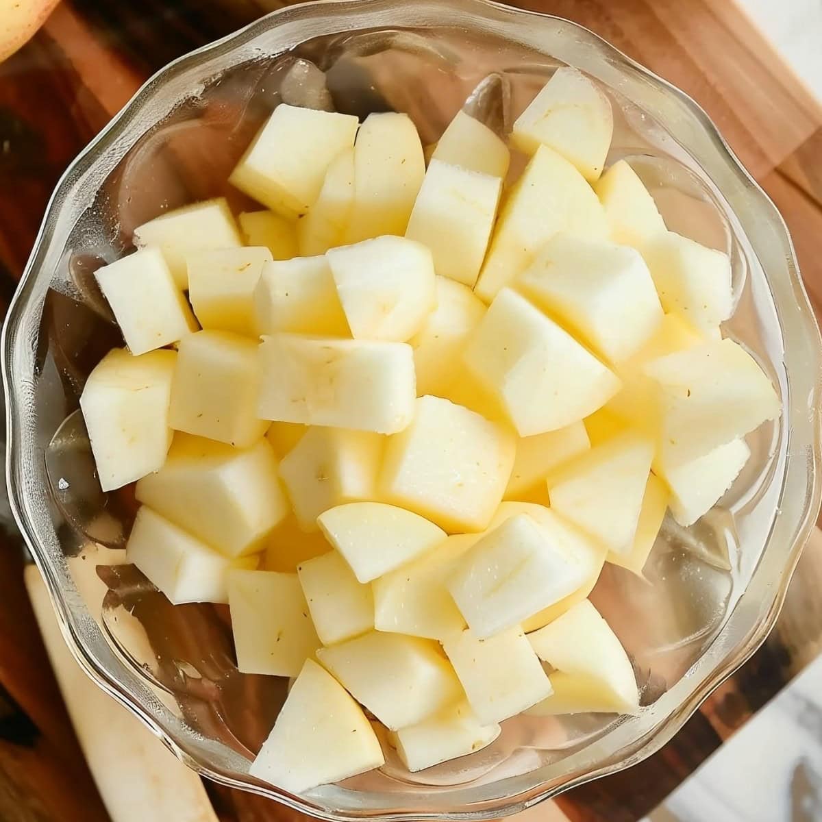 Top View of Peeled and Chopped Apple Pieces in a Glass Dish on a Wooden Cutting Board on a White Marble Table