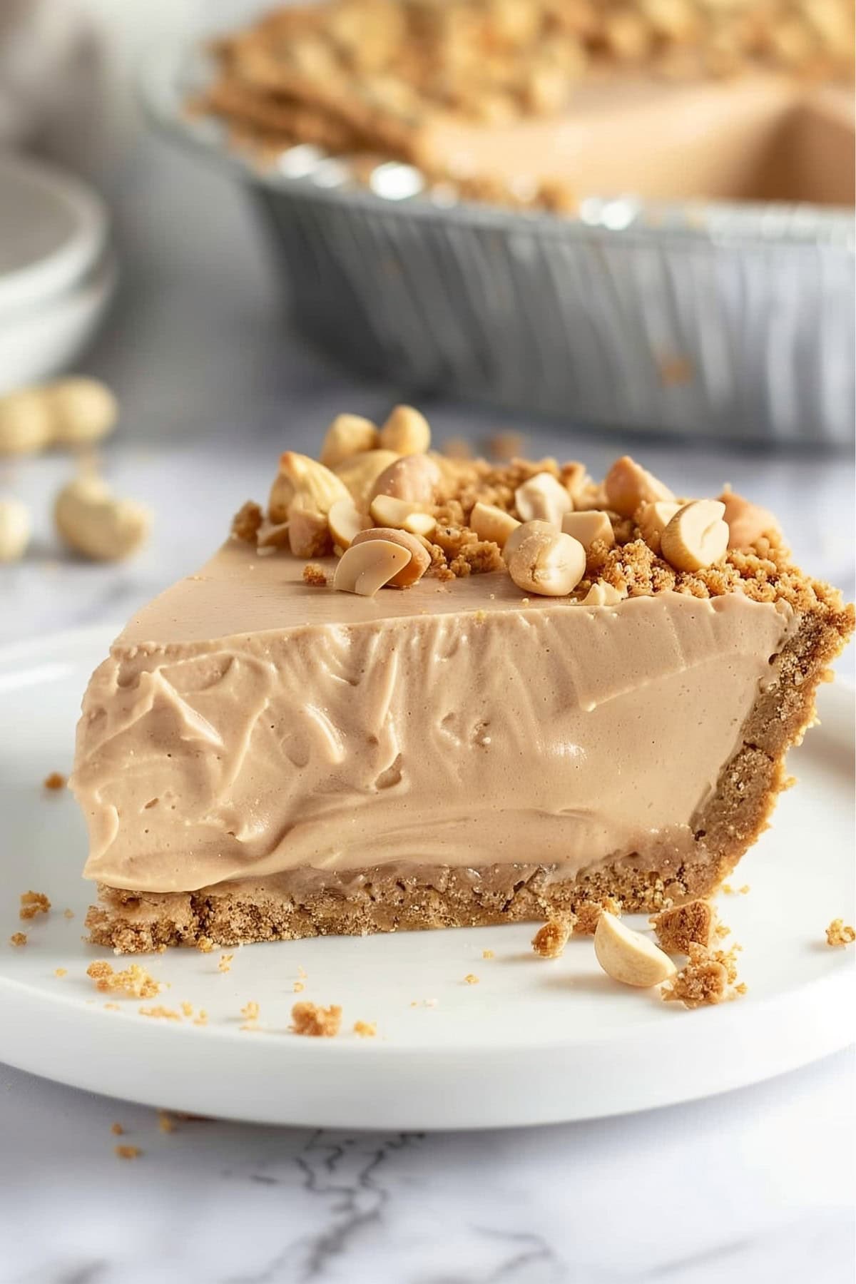 Close up of a Slice of Peanut Butter Cream Pie, Topped with Peanuts and Graham Cracker Crumbles on a White Plate with the Whole Pie in the Background