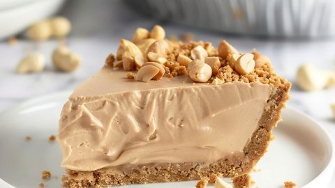 Close up of a Slice of Peanut Butter Cream Pie, Topped with Peanuts and Graham Cracker Crumbles on a White Plate with the Whole Pie in the Background