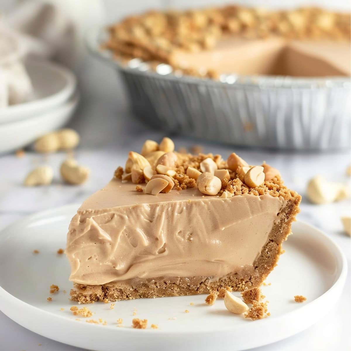 Close up of a Slice of Peanut Butter Cream Pie, Topped with Peanuts and Graham Cracker Crumbles on a White Plate with the Whole Pie in the Background