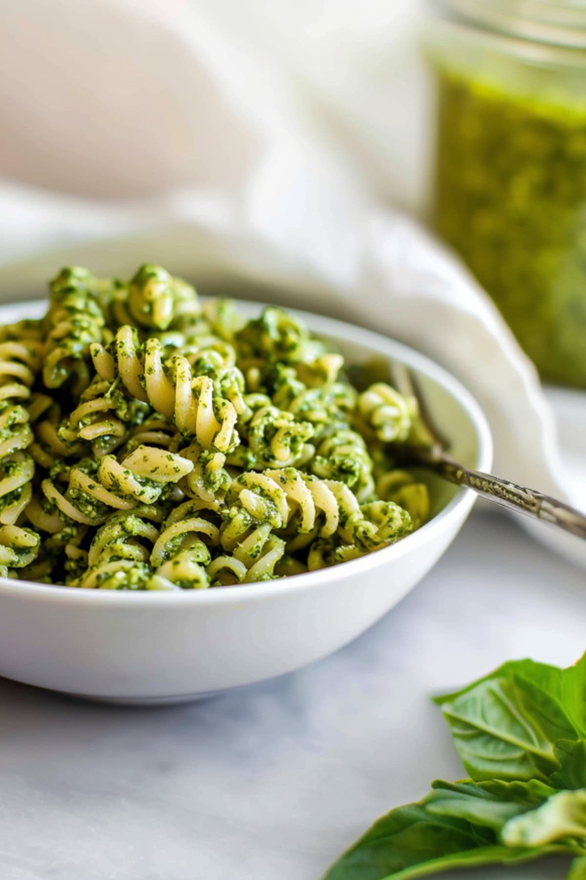 Side View Close Up of Pesto Pasta in a White Bowl with a Fork and a Jar of Green Basil Pesto in the Background