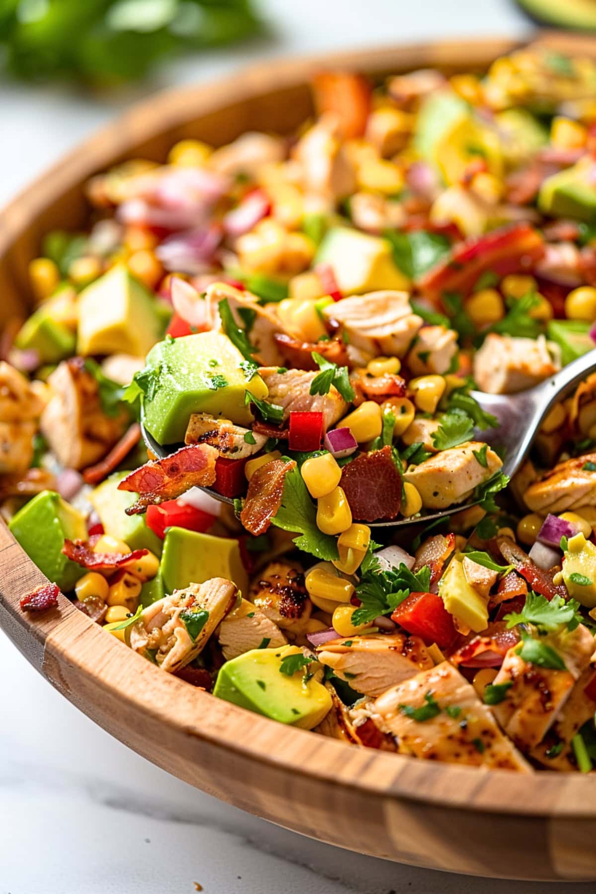 Spoon scooping avocado salad with with mix of bacon, cooked and crumbled, shredded chicken breasts, small square slice ripe avocados, corn kernels, finely chopped small red onion, fresh cilantro chopped with olive oil and lime juice dressing served in a wooden bowl.