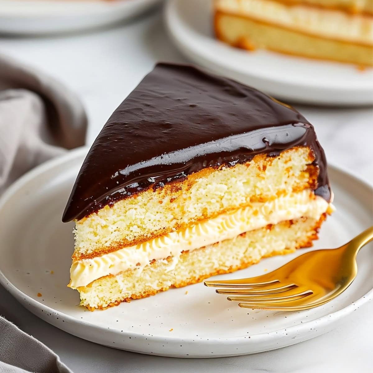 A slice of Boston cream pie on a white plate, showing layers of sponge cake and custard.