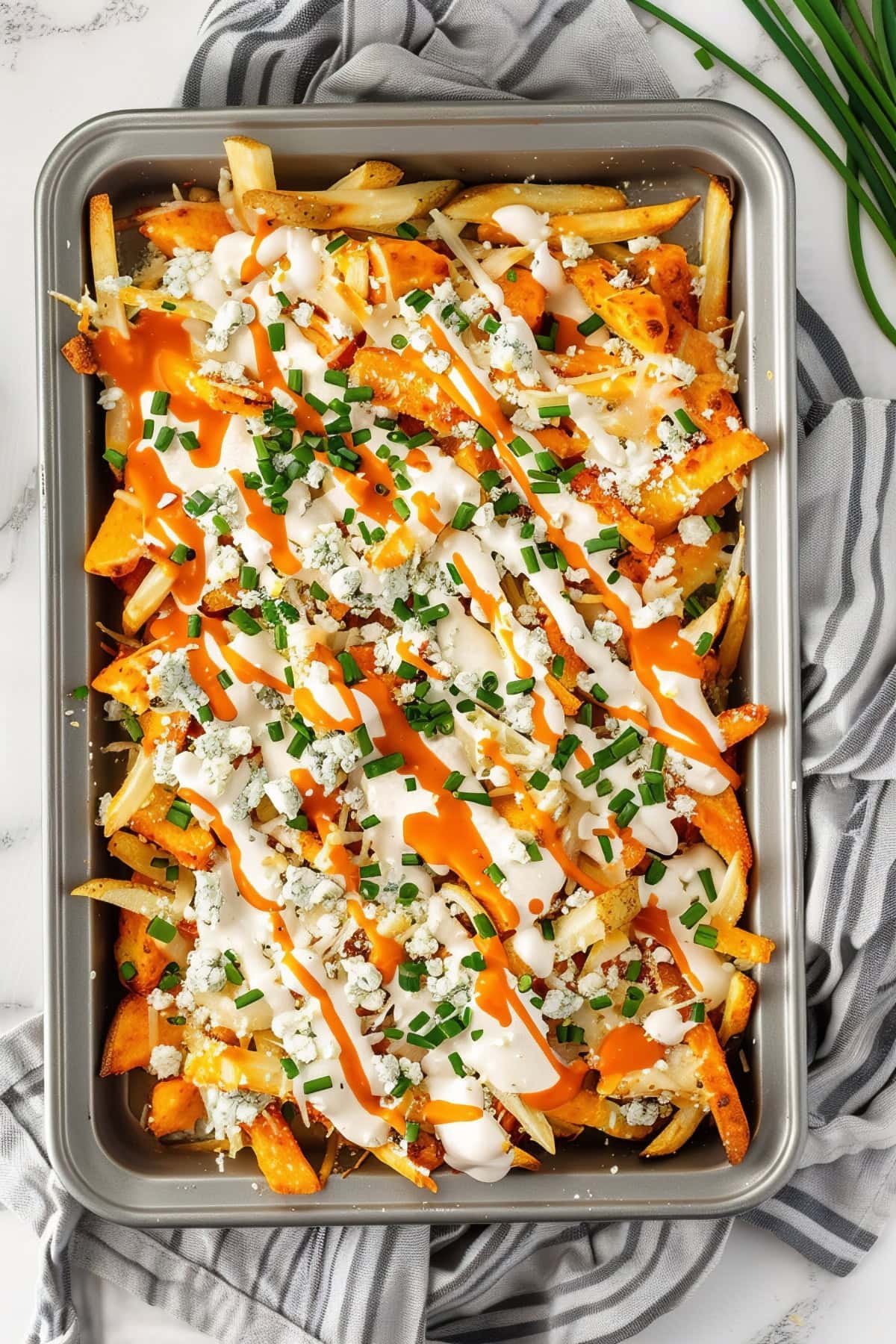 Fully loaded Buffalo fries topped with melted cheese, blue cheese crumbles, Buffalo sauce, ranch dressing, and chopped chives, on a sheet tray, top down view