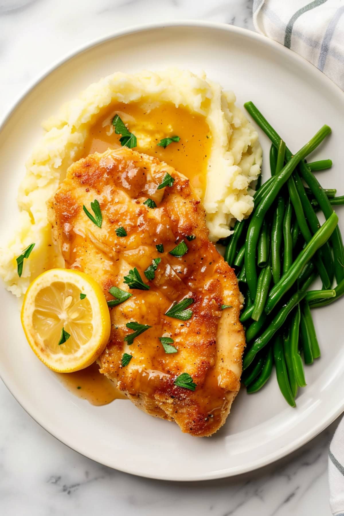 A plate full of chicken francese, green beans, mashed potatoes and a slice of lemon.