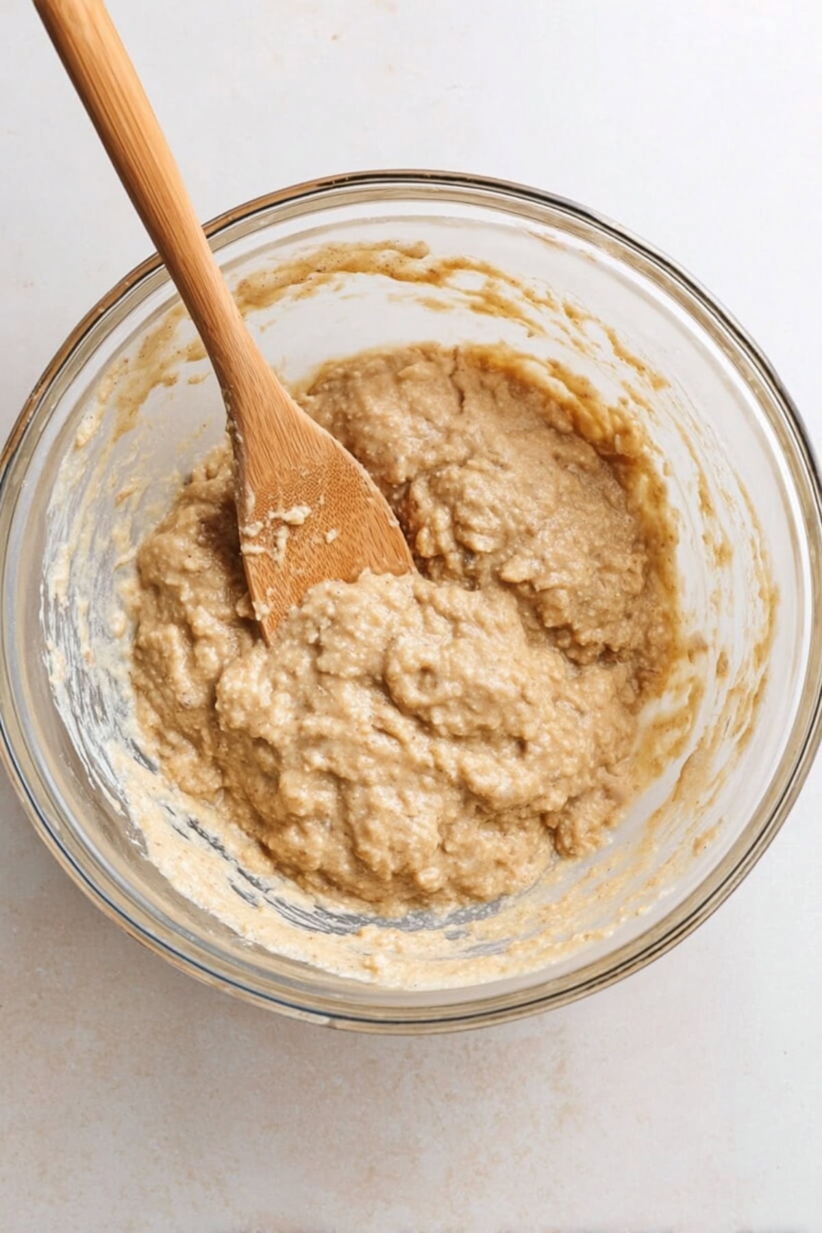 A glass mixing bowl filled with thick banana bread batter, being stirred with a wooden spoon.