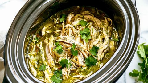 Salsa verde chicken in a slow cooker sitting on a white marble table.