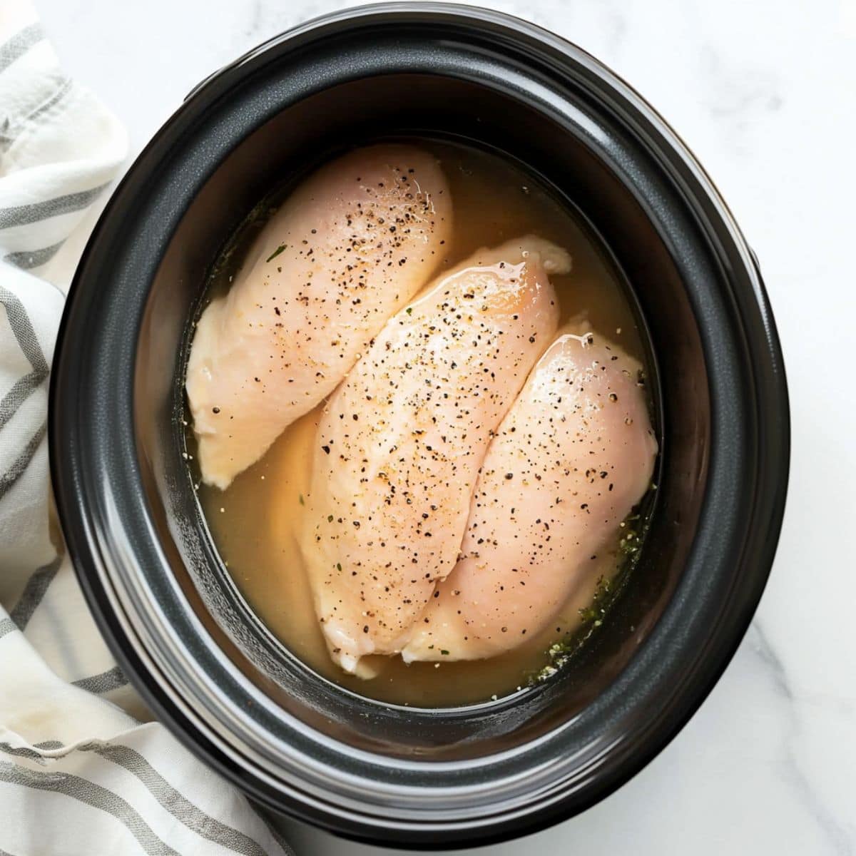 Seasoned raw chicken breast in a slow cooker pot with broth.