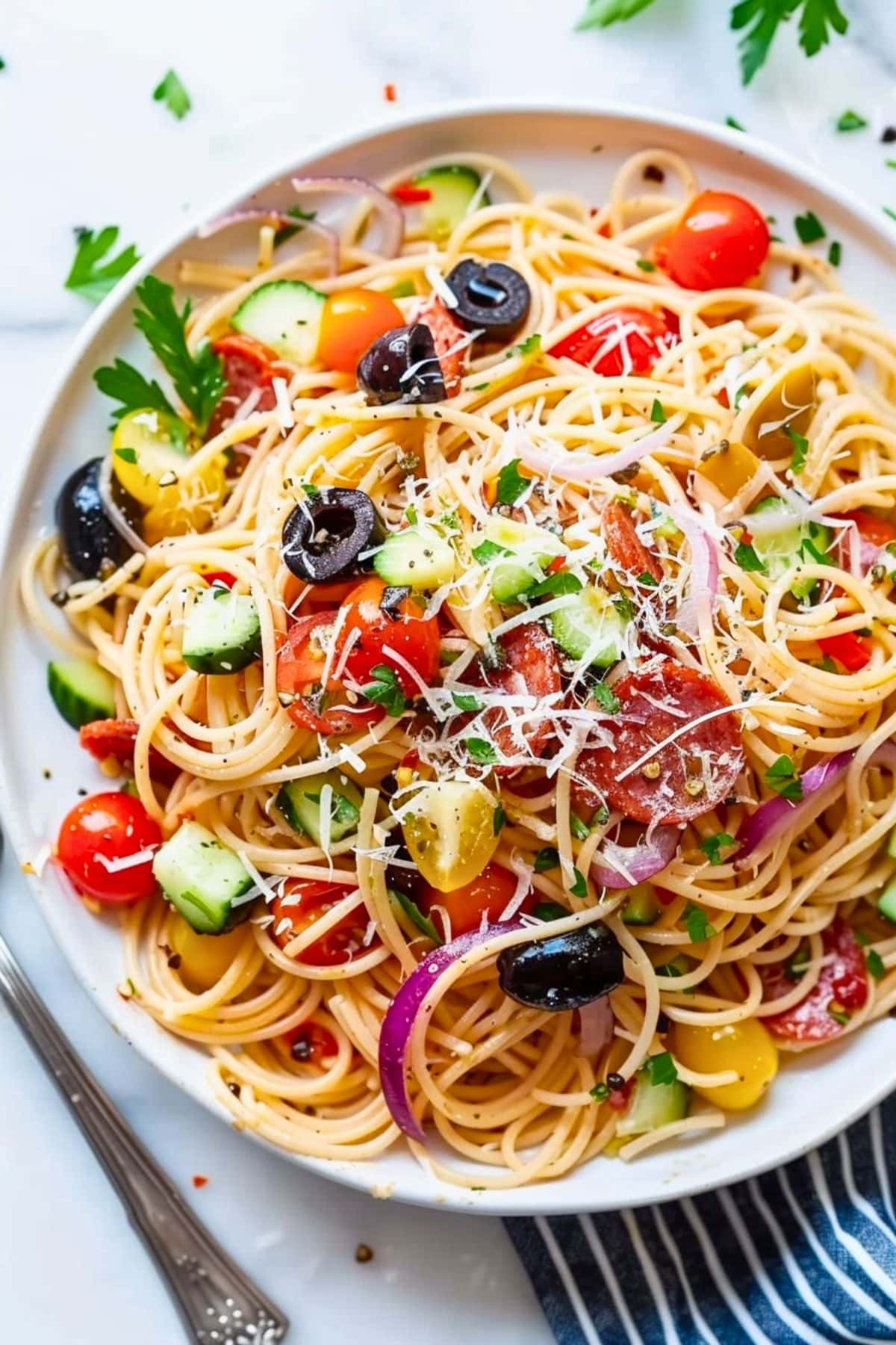 Italian spaghetti pasta made with made with thin spaghetti broken into thirds, grated Parmesan cheese, seeded and chopped red bell pepper, thinly sliced red onion, halved cherry tomatoes, and mini pepperoni, sliced black olives served in a white plate, fork on the side.