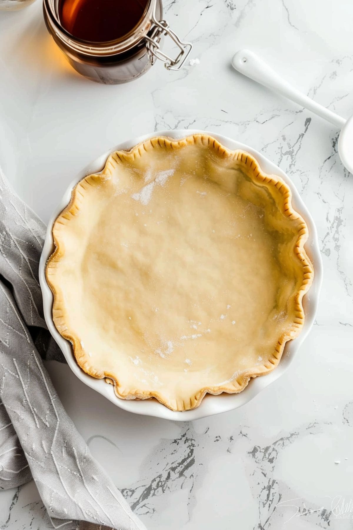 Pie crust in a white pie dish and a jar with dark maple syrup next to it.