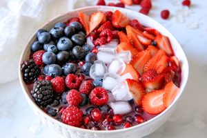 Mix of blueberries, blackberries, raspberries, pomegranate seeds, and chopped strawberries in a white bowl with cube ice and coconut water.