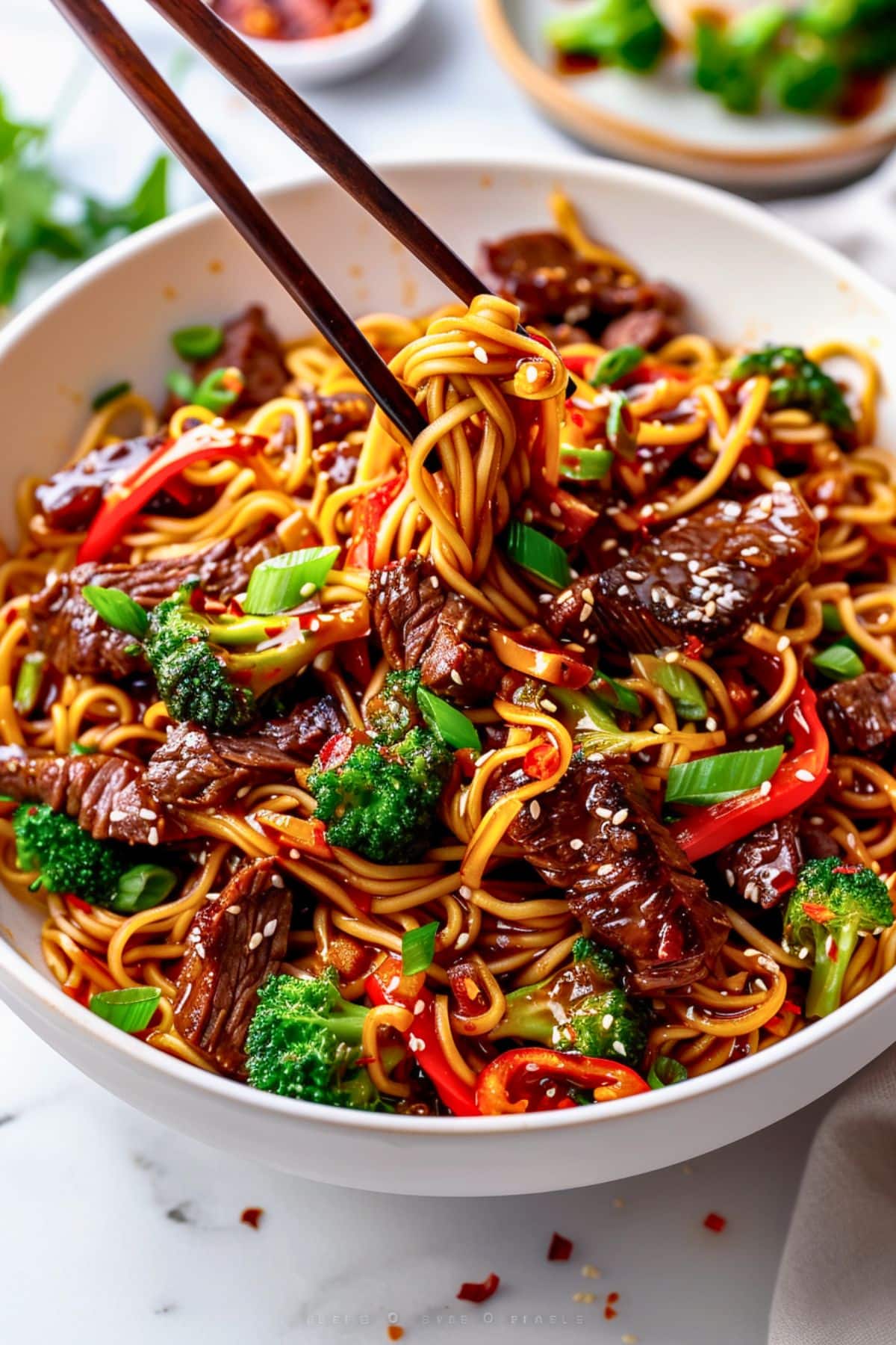 Chopsticks lifting noodles from a bowl serving of sticky beef noodles. 