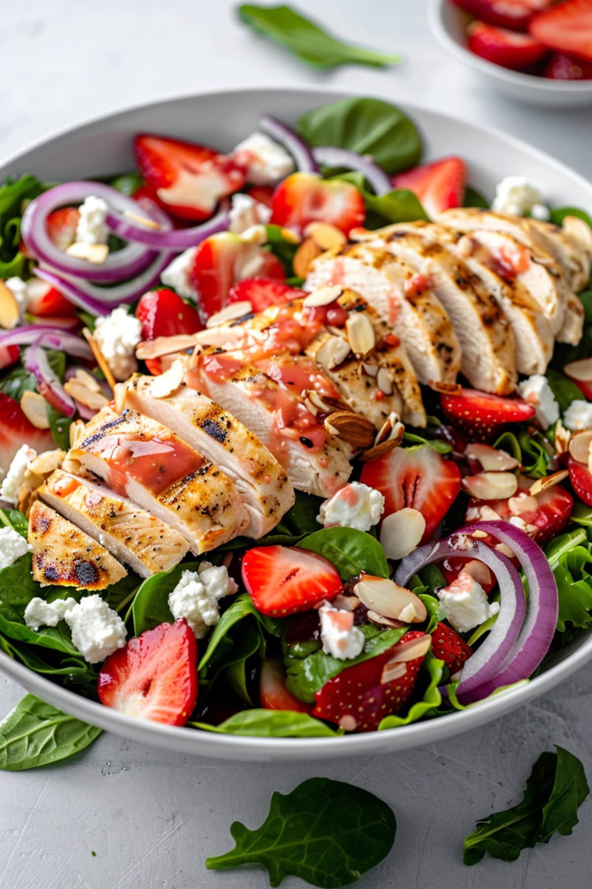 A serving of salad with mixed greens, feta cheese, sliced almonds and onions topped with sliced grilled chicken breast drizzled with strawberry dressing on a white bowl.
