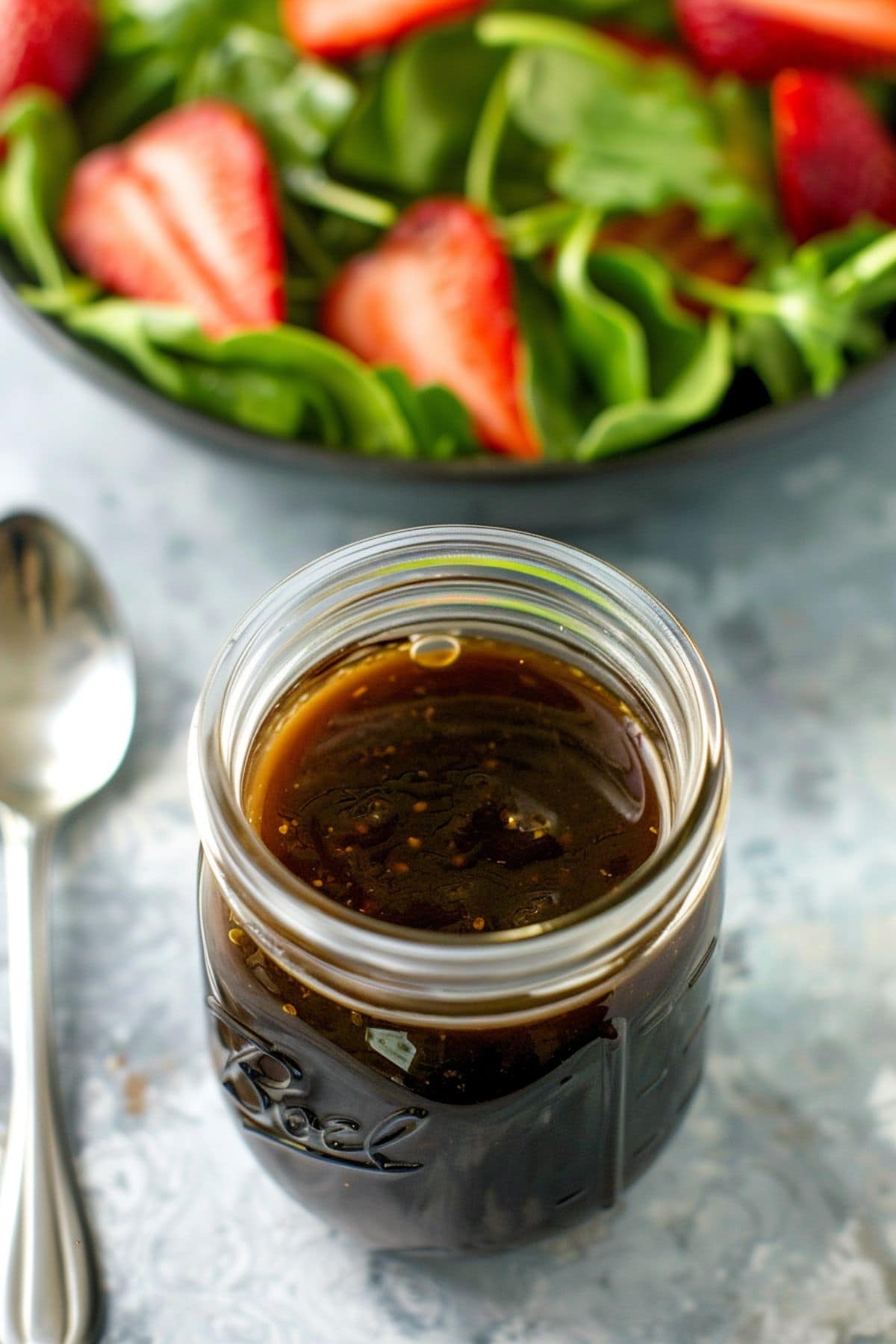 Balsamic dressing in a small glass jar, bowl of strawberry salad sitting next to it.