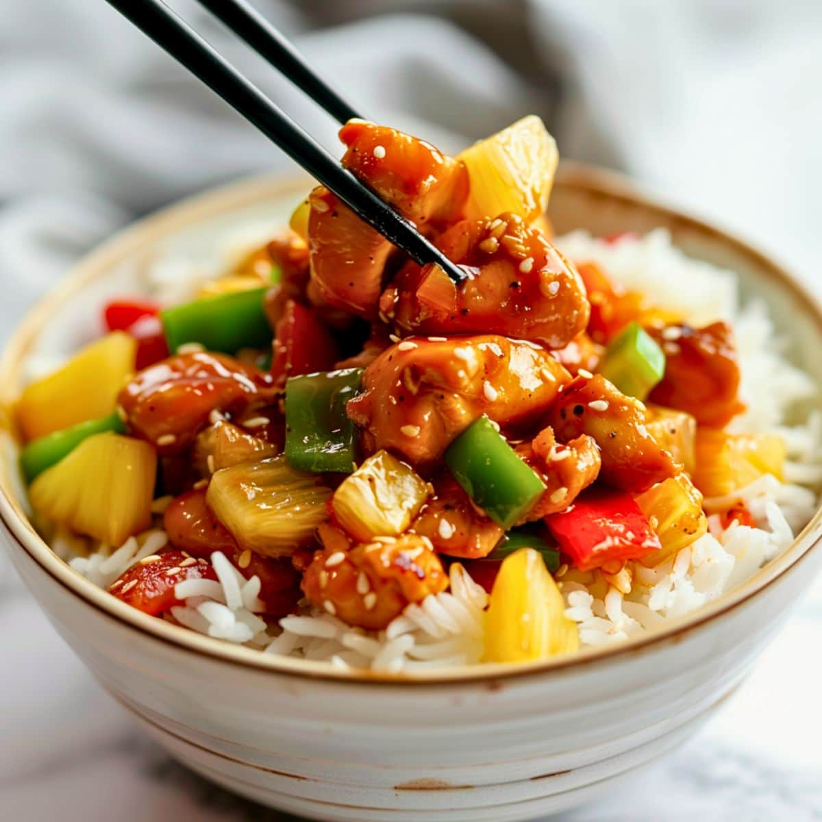 Chopsticks lifting a piece of chicken topped from a bowl of white rice with pineapple chunks, green and red bell pepper.