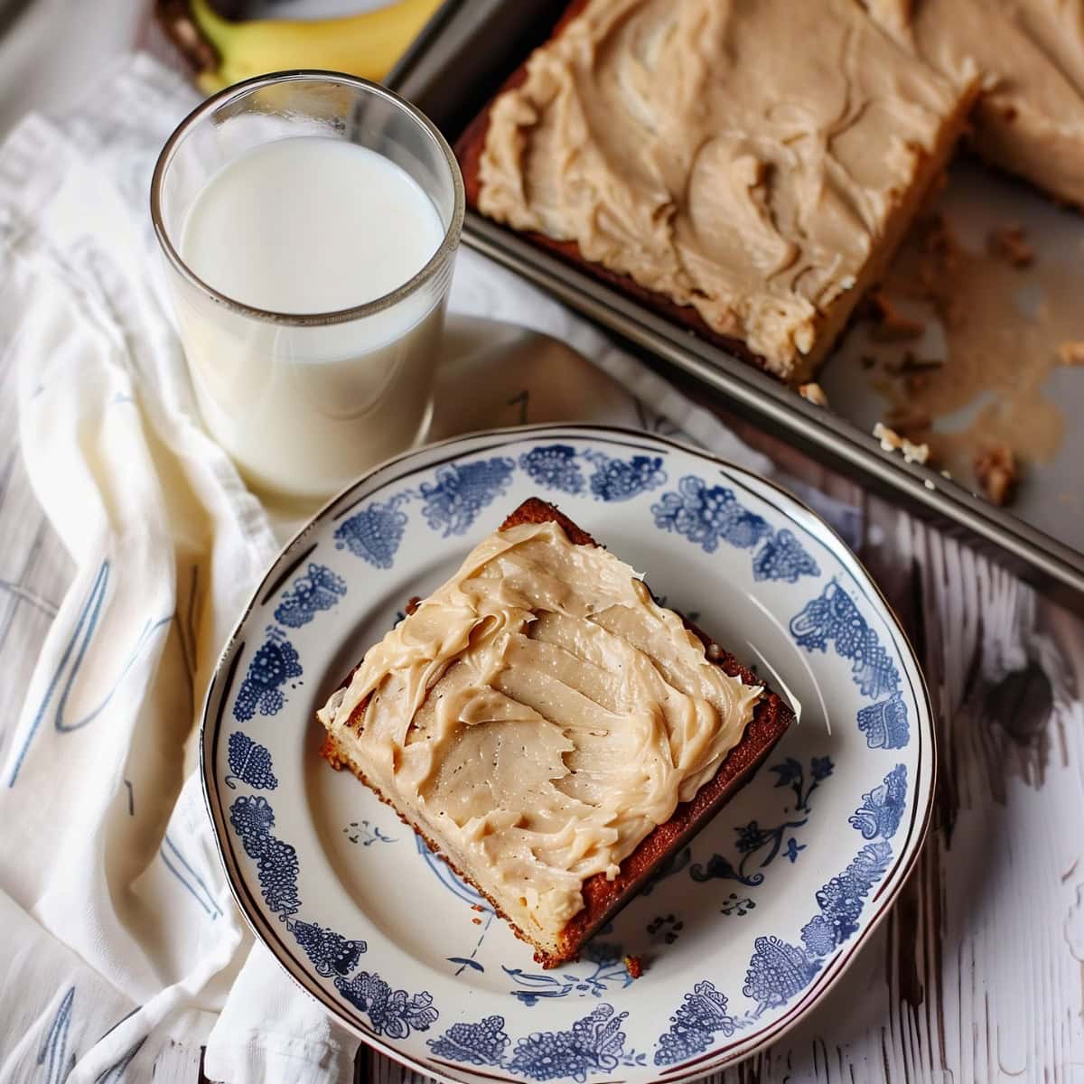 Top view of a slice of banana bread with brown butter frosting served on a  plate and a glass of milk next to it.